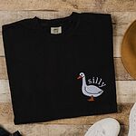 Black T-shirt laid flat on wood beside white sneakers, embroidered with a white goose and the word silly in white thread