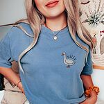 Woman in a blue T-shirt and a gold necklace, embroidered with a small white goose and the word silly
