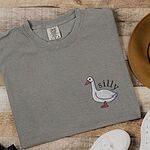 Grey T-shirt laid flat on wood beside a felt hat and sneakers, embroidered with a little white goose with the word silly arched over its back on the left chest