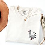 Embroidered goose shirt in Ivory laid flat with a gold necklace and a felt hat, stitched with a little white goose with the word silly arched over its back on the left chest