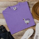 Violet T-shirt laid flat on wood with jeans and sneakers, embroidered with a little white goose with the word silly arched over its back on the left chest