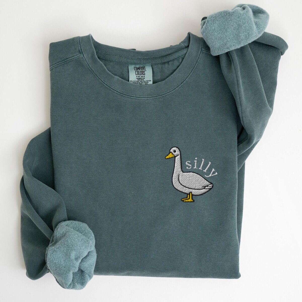 Embroidered Goose Sweatshirt – Blue Spruce Blue Spruce crewneck sweatshirt laid flat, embroidered with a white goose and the word silly in white thread on the left chest
