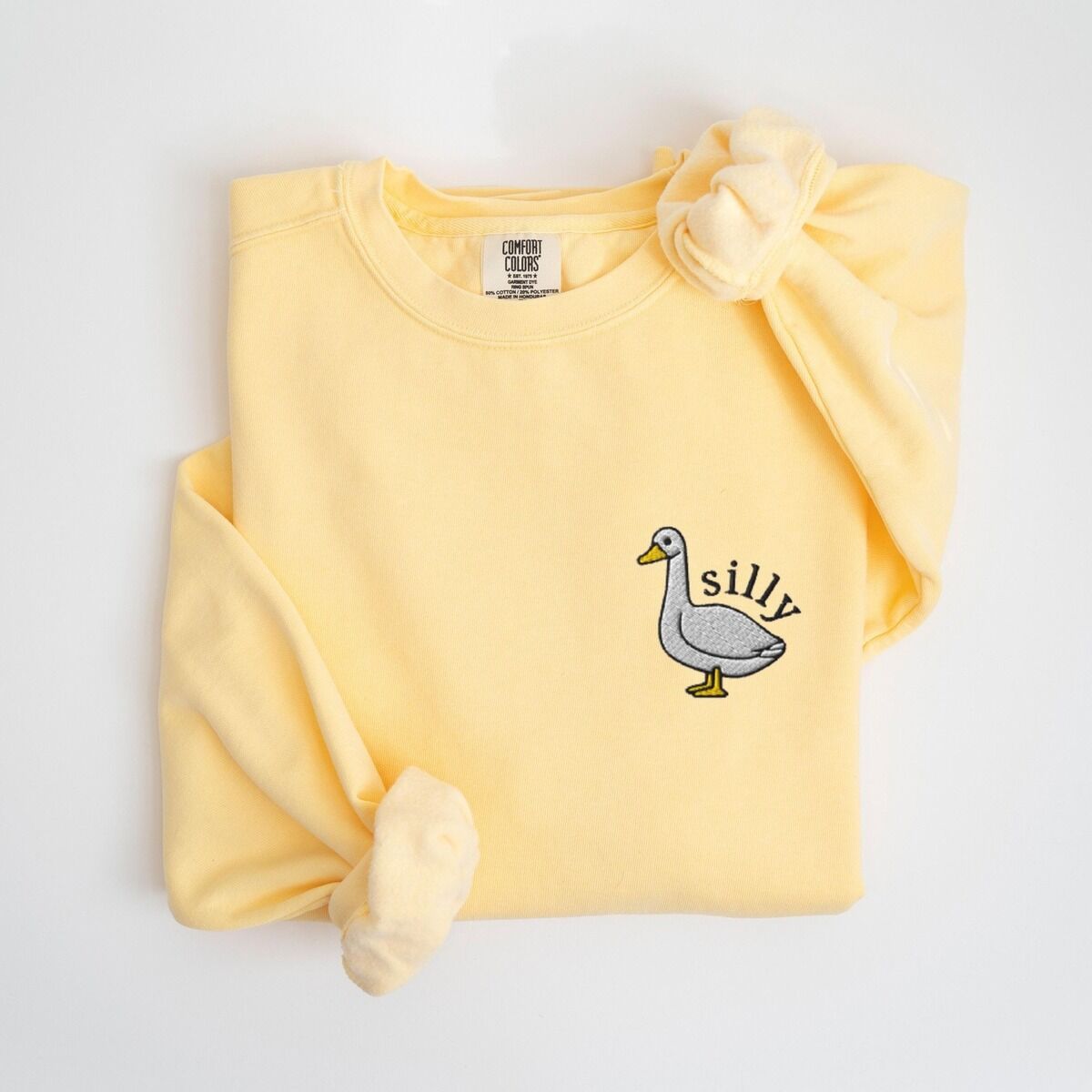 Embroidered Goose Sweatshirt – Butter Butter yellow crewneck sweatshirt laid flat with a knotted sleeve, embroidered with a little white goose with the word silly arched over its back on the left chest