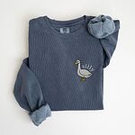 Denim crewneck sweatshirt laid flat with rolled cuffs, embroidered with a white goose and the word silly in white thread on the left chest