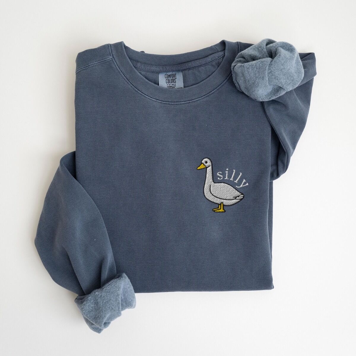 Embroidered Goose Sweatshirt – Denim Denim crewneck sweatshirt laid flat with rolled cuffs, embroidered with a white goose and the word silly in white thread on the left chest