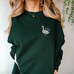 Close view of a woman in a forest green crewneck sweatshirt embroidered with a white goose and the word silly
