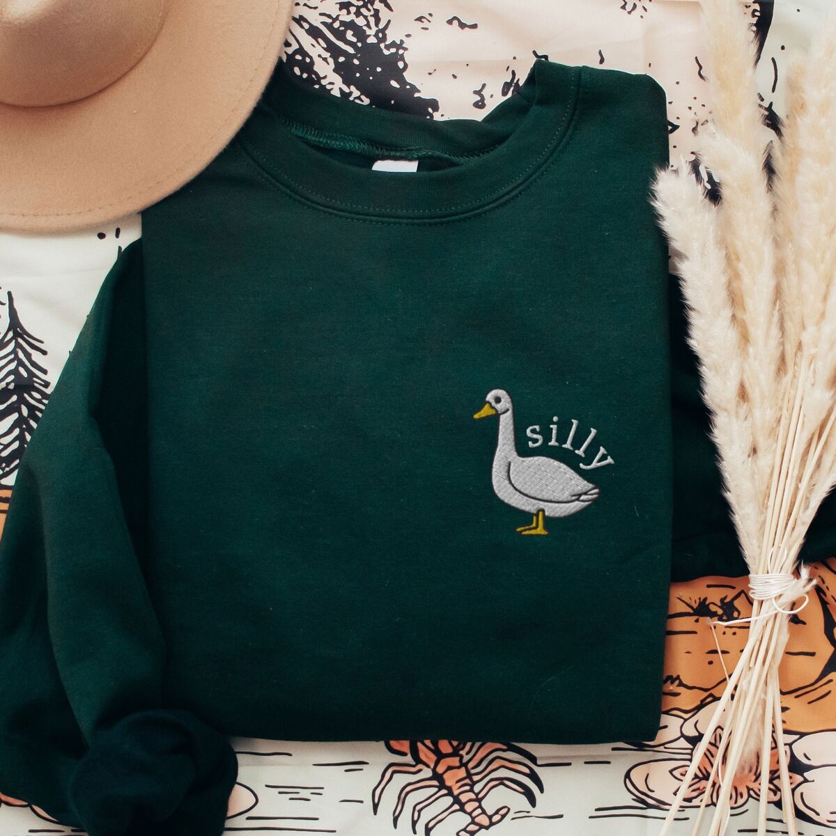 Embroidered Goose Sweatshirt - Forest Green Forest Green crewneck sweatshirt laid flat on a printed blanket with dried pampas grass, embroidered with a white goose and the word silly in white thread