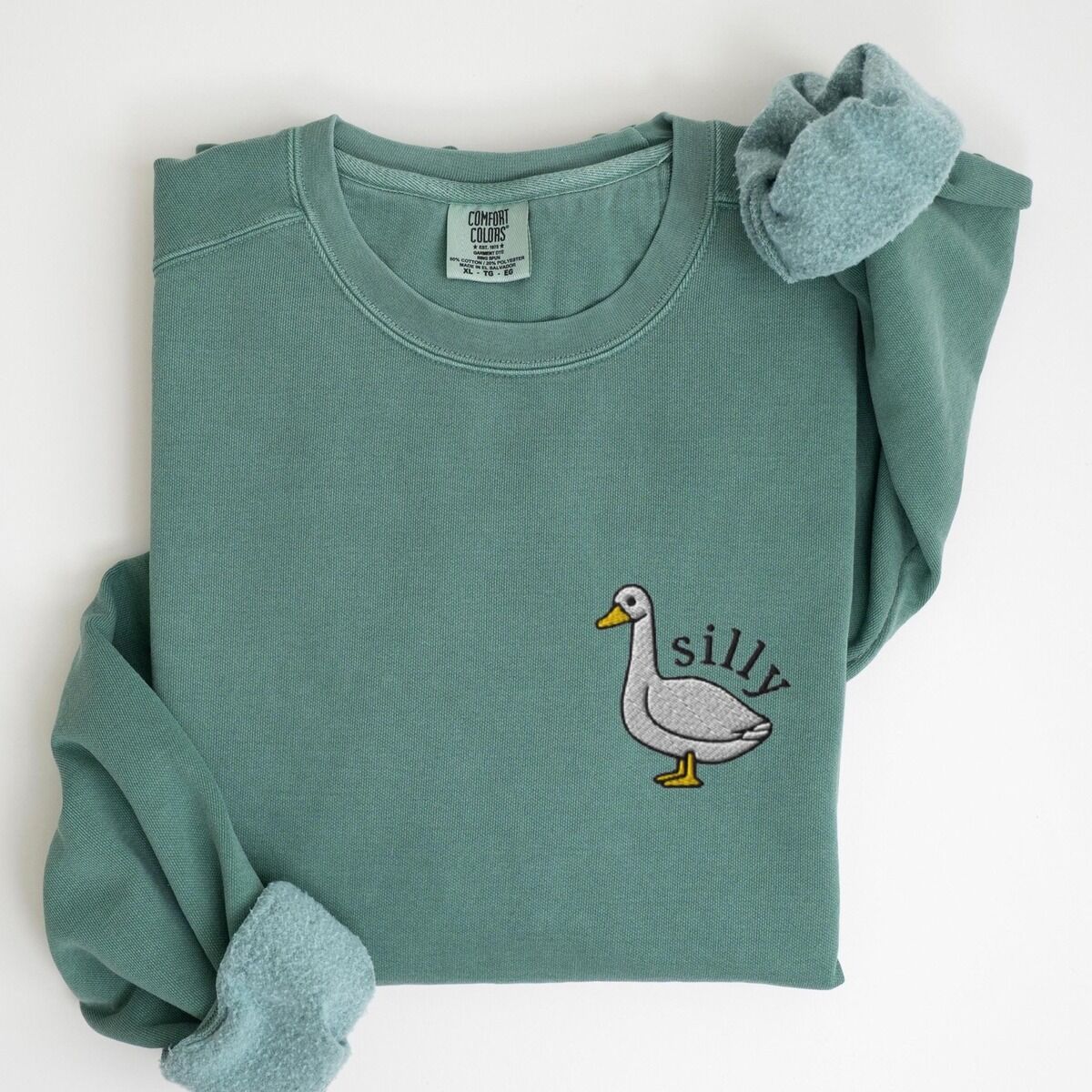 Embroidered Goose Sweatshirt – Light Green Embroidered goose sweatshirt in Light Green laid flat with rolled cuffs, stitched with a little white goose with the word silly arched over its back on the left chest