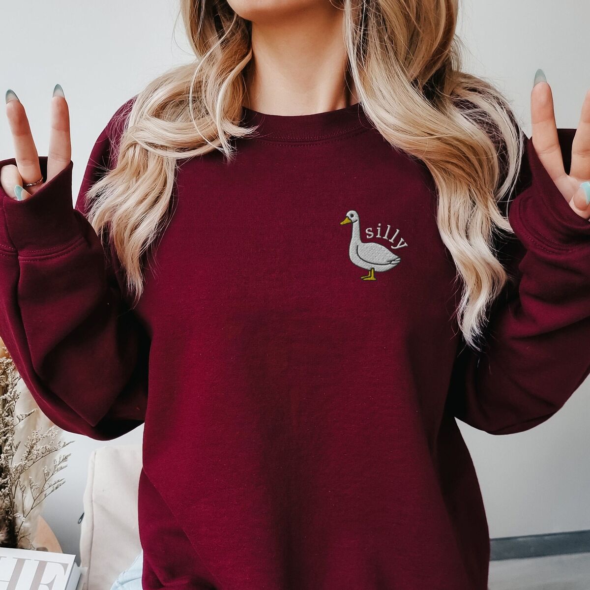 Embroidered Goose Sweatshirt - Maroon Woman making peace signs in a Maroon crewneck sweatshirt embroidered with a white goose and the word silly