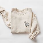 Sand crewneck sweatshirt laid flat with a rolled cuff, embroidered with a little white goose with the word silly arched over its back on the left chest