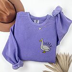 Violet crewneck sweatshirt laid flat beside a felt hat and a dried palm leaf, embroidered with a little white goose with the word silly arched over its back on the left chest