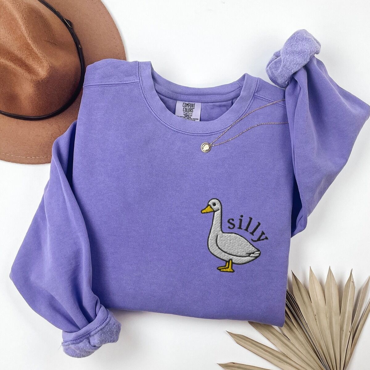 Embroidered Goose Sweatshirt – Violet Violet crewneck sweatshirt laid flat beside a felt hat and a dried palm leaf, embroidered with a little white goose with the word silly arched over its back on the left chest