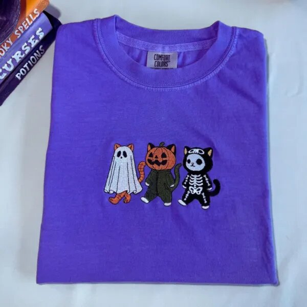 Violet T-shirt with an embroidered ghost cat, pumpkin cat and skeleton cat across the chest