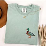 Embroidered mallard shirt in Bay laid flat with a gold necklace and a felt hat, stitched with a standing drake mallard with a green head, orange bill, chestnut chest and grey wing on the left chest