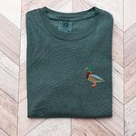 Blue Spruce T-shirt folded on a chevron wood floor, embroidered with a standing drake mallard with a green head, orange bill, chestnut chest and grey wing on the left chest