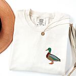 Ivory T-shirt laid flat with a coin necklace and a dried palm leaf, embroidered with a standing drake mallard with a green head, orange bill, chestnut chest and grey wing on the left chest