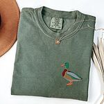 Moss T-shirt laid flat with a gold necklace, embroidered with a standing drake mallard with a green head, orange bill, chestnut chest and grey wing on the left chest