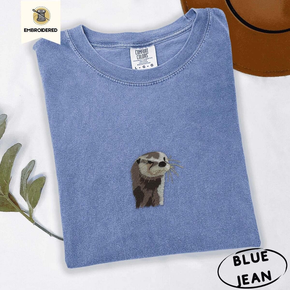 Embroidered Otter Shirt – Blue Jean Blue Jean Comfort Colors T-shirt folded beside a brown felt hat and a eucalyptus sprig, embroidered with a river otter head in brown and cream thread with long golden whiskers