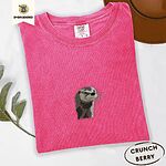 Crunchberry Comfort Colors T-shirt folded beside a brown felt hat and a eucalyptus sprig, embroidered with a river otter head in brown and cream thread with long golden whiskers