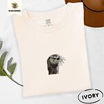 Ivory Comfort Colors T-shirt folded beside a brown felt hat and a eucalyptus sprig, embroidered with a river otter head in brown and cream thread with long golden whiskers