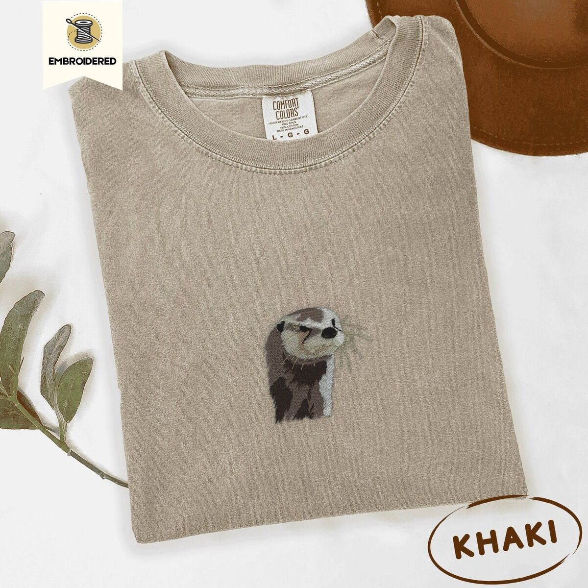 Embroidered Otter Shirt – Khaki Khaki Comfort Colors T-shirt folded beside a brown felt hat and a eucalyptus sprig, embroidered with a river otter head in brown and cream thread with long golden whiskers