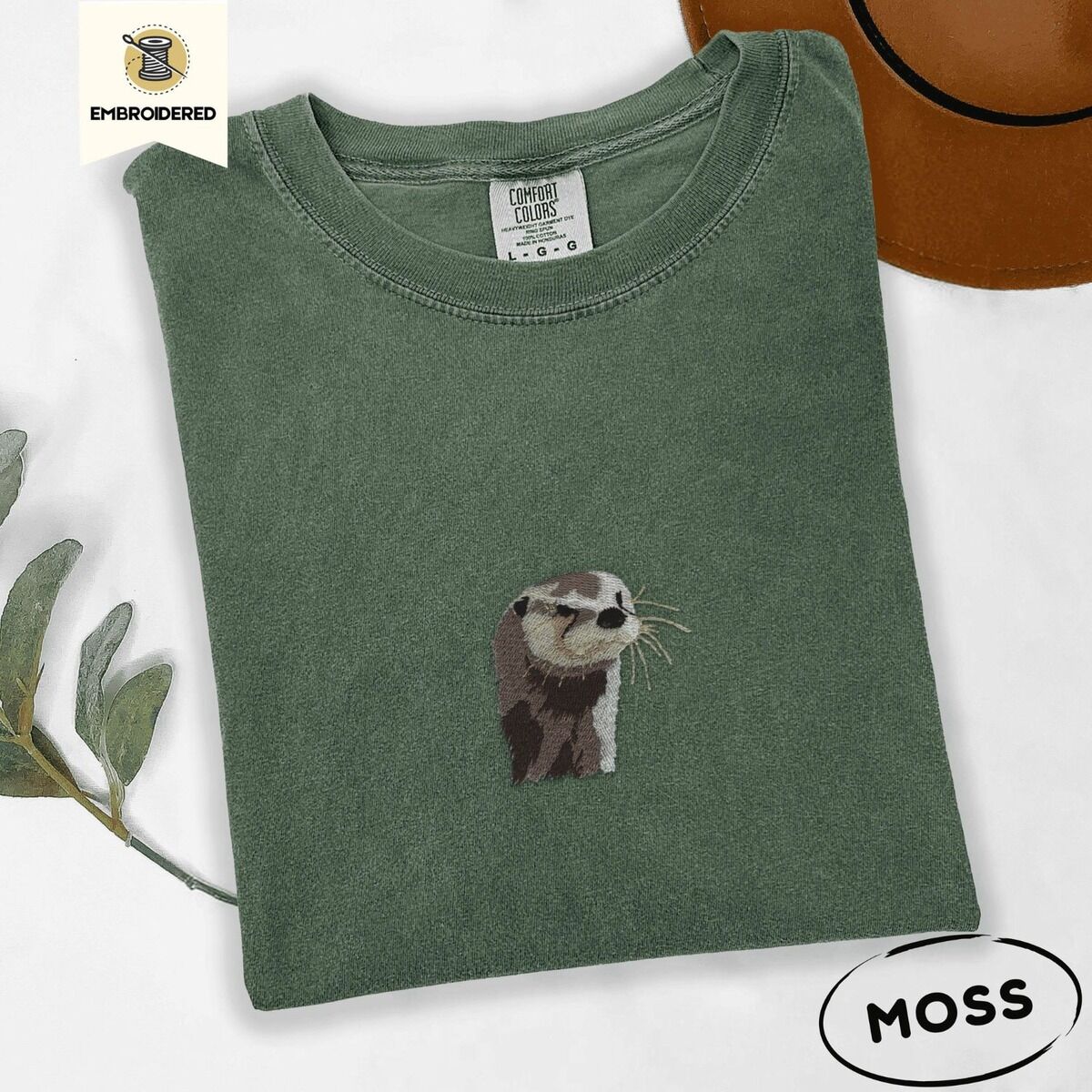 Embroidered Otter Shirt – Moss Moss Comfort Colors T-shirt folded beside a brown felt hat and a eucalyptus sprig, embroidered with a river otter head in brown and cream thread with long golden whiskers