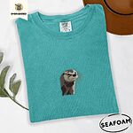 Seafoam Comfort Colors T-shirt folded beside a brown felt hat and a eucalyptus sprig, embroidered with a river otter head in brown and cream thread with long golden whiskers