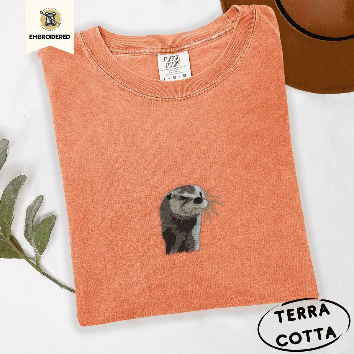 Embroidered Otter Shirt – Terracotta Terracotta Comfort Colors T-shirt folded beside a brown felt hat and a eucalyptus sprig, embroidered with a river otter head in brown and cream thread with long golden whiskers
