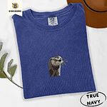 True Navy Comfort Colors T-shirt folded beside a brown felt hat and a eucalyptus sprig, embroidered with a river otter head in brown and cream thread with long golden whiskers