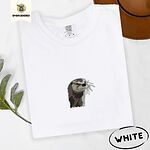 White Comfort Colors T-shirt folded beside a brown felt hat and a eucalyptus sprig, embroidered with a river otter head in brown and cream thread with long golden whiskers