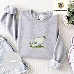 Ash crewneck sweatshirt folded with denim shorts, white sneakers and a potted plant, embroidered with a woolly white ewe standing over her lamb resting on a patch of green grass