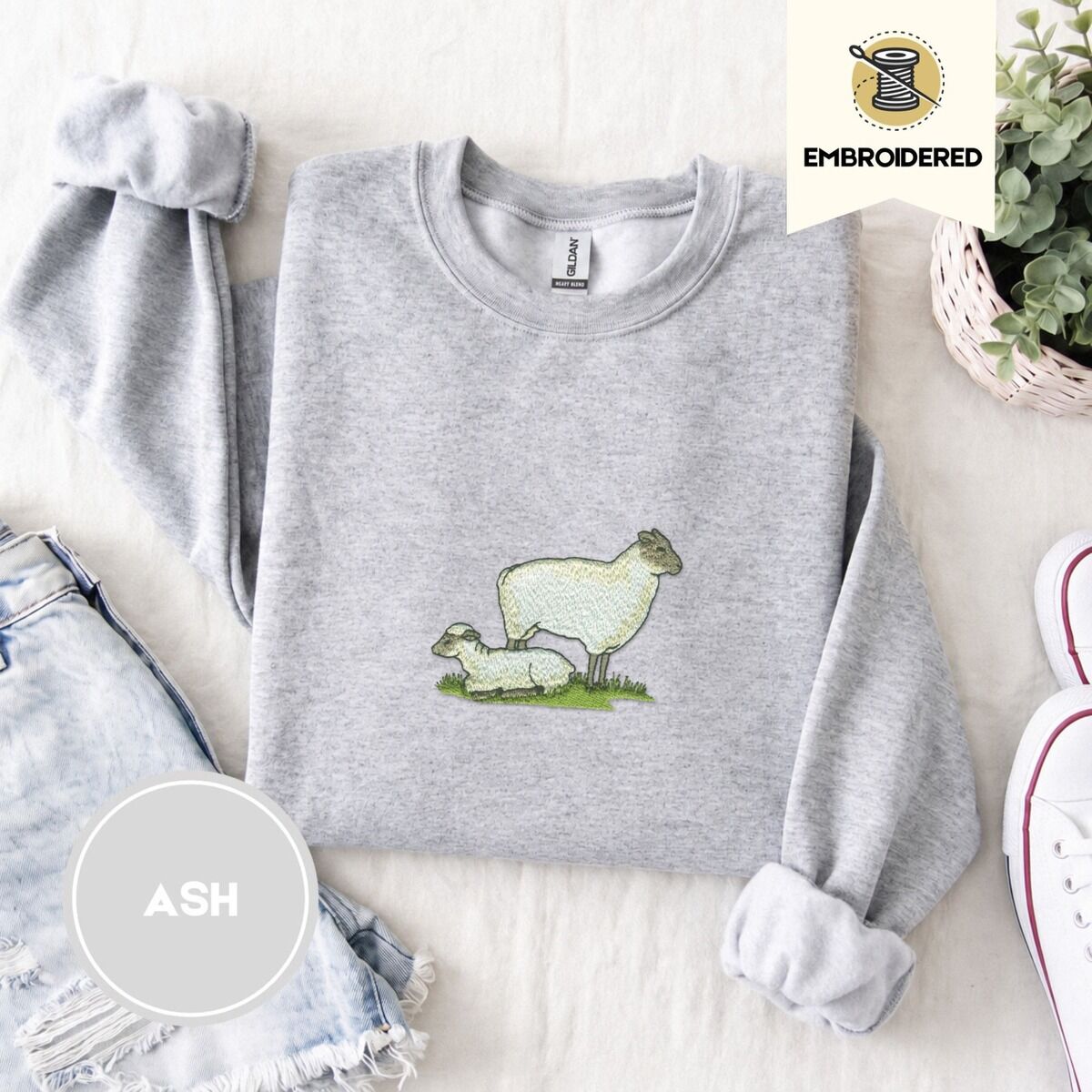 Embroidered Sheep Sweatshirt – Ash Ash crewneck sweatshirt folded with denim shorts, white sneakers and a potted plant, embroidered with a woolly white ewe standing over her lamb resting on a patch of green grass