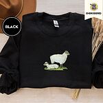 Black crewneck sweatshirt with a brown felt hat and dried bunny tail grass, embroidered with a woolly white ewe standing over her lamb resting on a patch of green grass