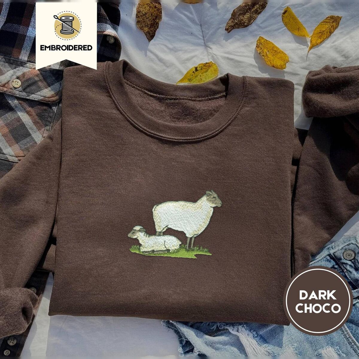 Embroidered Sheep Sweatshirt – Dark Chocolate Dark Chocolate crewneck sweatshirt on a plaid flannel shirt with yellow autumn leaves and denim shorts, embroidered with a woolly white ewe standing over her lamb resting on a patch of green grass