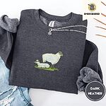 Dark Heather crewneck sweatshirt folded with ripped jeans and a silver chain necklace, embroidered with a woolly white ewe standing over her lamb resting on a patch of green grass