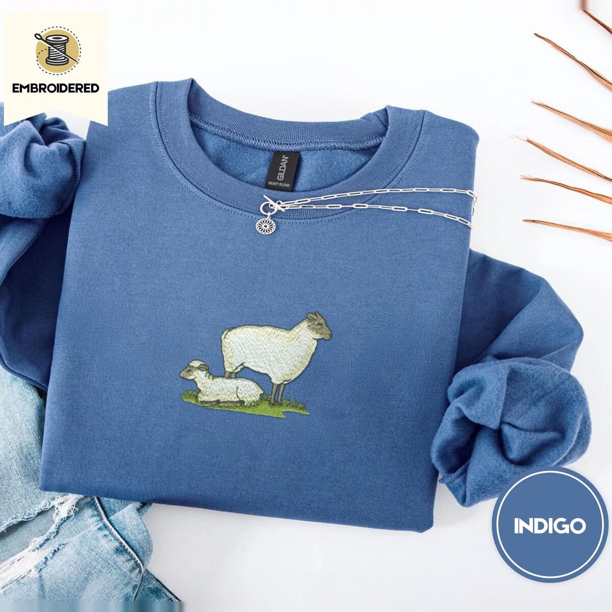 Embroidered Sheep Sweatshirt – Indigo Blue Indigo Blue crewneck sweatshirt folded with light jeans and dried twigs, embroidered with a woolly white ewe standing over her lamb resting on a patch of green grass