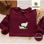 Maroon crewneck sweatshirt with a brown felt hat and dried bunny tail grass, embroidered with a woolly white ewe standing over her lamb resting on a patch of green grass