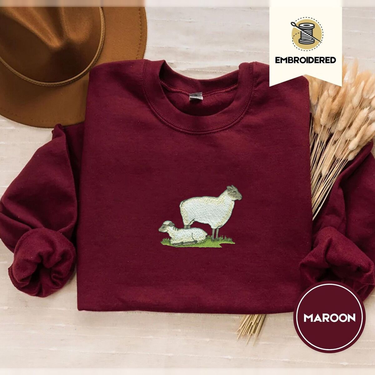 Embroidered Sheep Sweatshirt – Maroon Maroon crewneck sweatshirt with a brown felt hat and dried bunny tail grass, embroidered with a woolly white ewe standing over her lamb resting on a patch of green grass