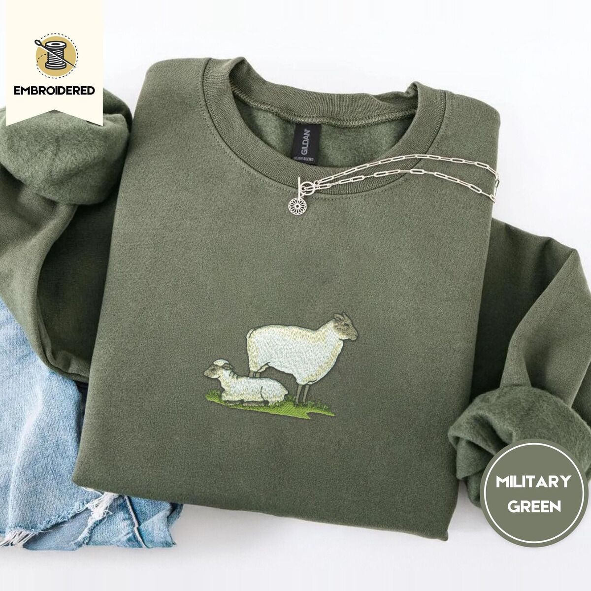Embroidered Sheep Sweatshirt – Military Green Military Green crewneck sweatshirt folded with light jeans and a silver chain necklace, embroidered with a woolly white ewe standing over her lamb resting on a patch of green grass