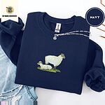 Navy crewneck sweatshirt folded with ripped jeans and a silver chain necklace, embroidered with a woolly white ewe standing over her lamb resting on a patch of green grass
