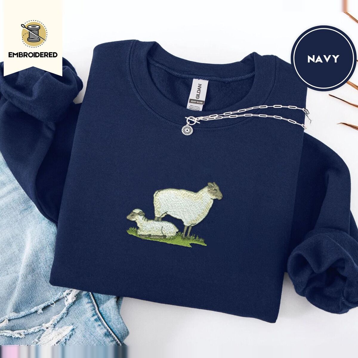 Embroidered Sheep Sweatshirt – Navy Navy crewneck sweatshirt folded with ripped jeans and a silver chain necklace, embroidered with a woolly white ewe standing over her lamb resting on a patch of green grass