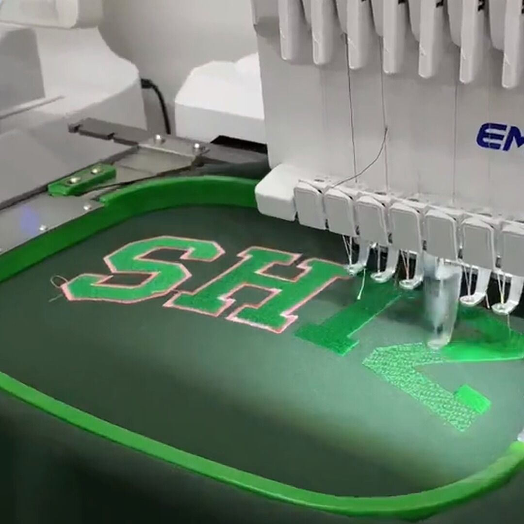 Our Embroidery Workshop – Video Cover Multi-needle embroidery machine stitching green varsity letters on a sweatshirt, from our workshop video