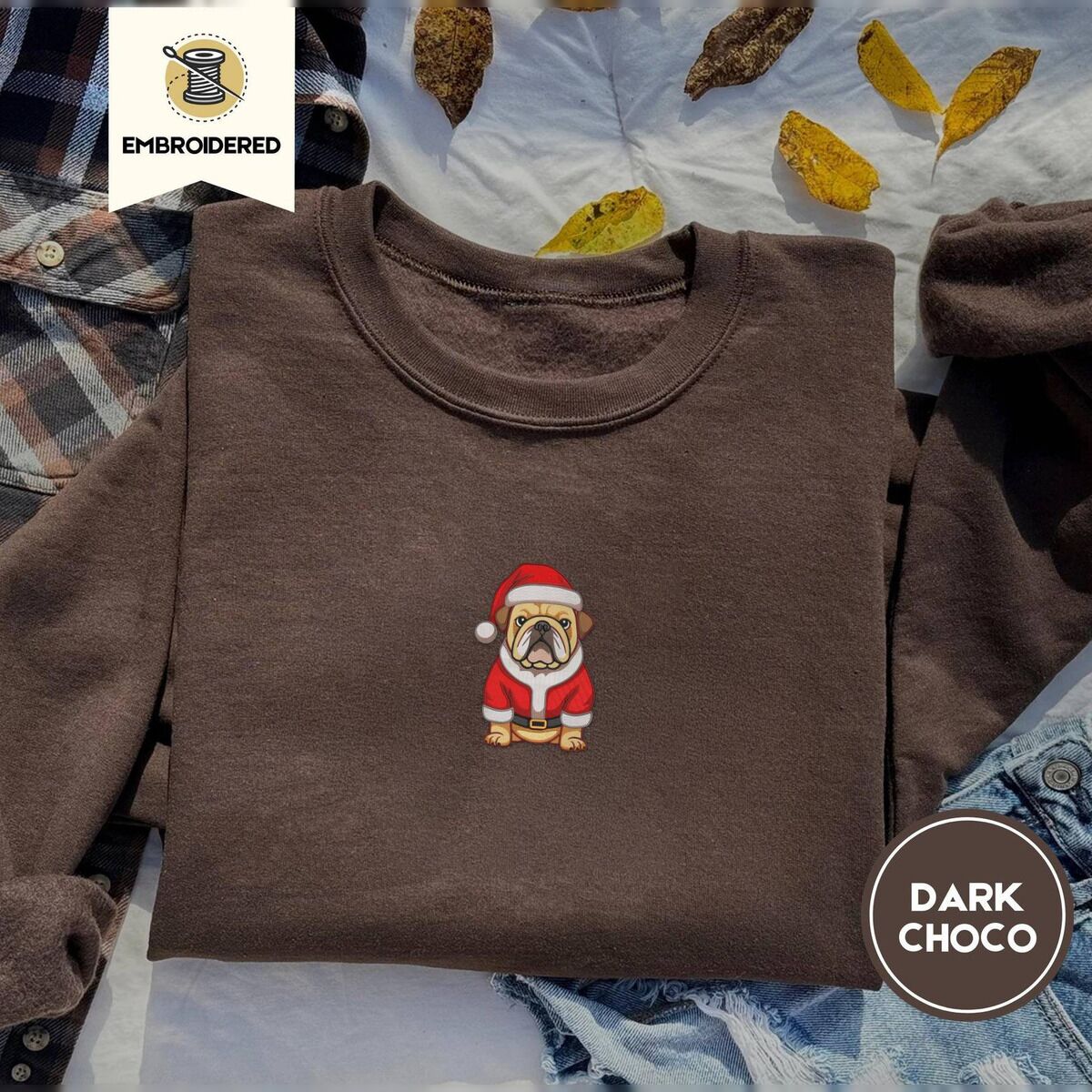 English Bulldog Christmas Sweatshirt – Dark Chocolate Dark Chocolate crewneck sweatshirt on a plaid flannel shirt with yellow autumn leaves and denim shorts, embroidered with a fawn English bulldog in a red Santa suit, black belt and Santa hat