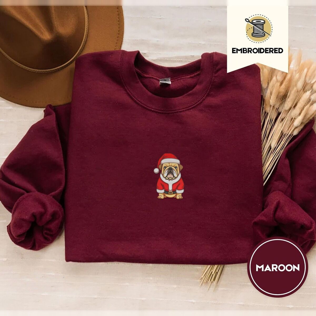 English Bulldog Christmas Sweatshirt – Maroon Maroon crewneck sweatshirt with a brown felt hat and dried bunny tail grass, embroidered with a fawn English bulldog in a red Santa suit, black belt and Santa hat