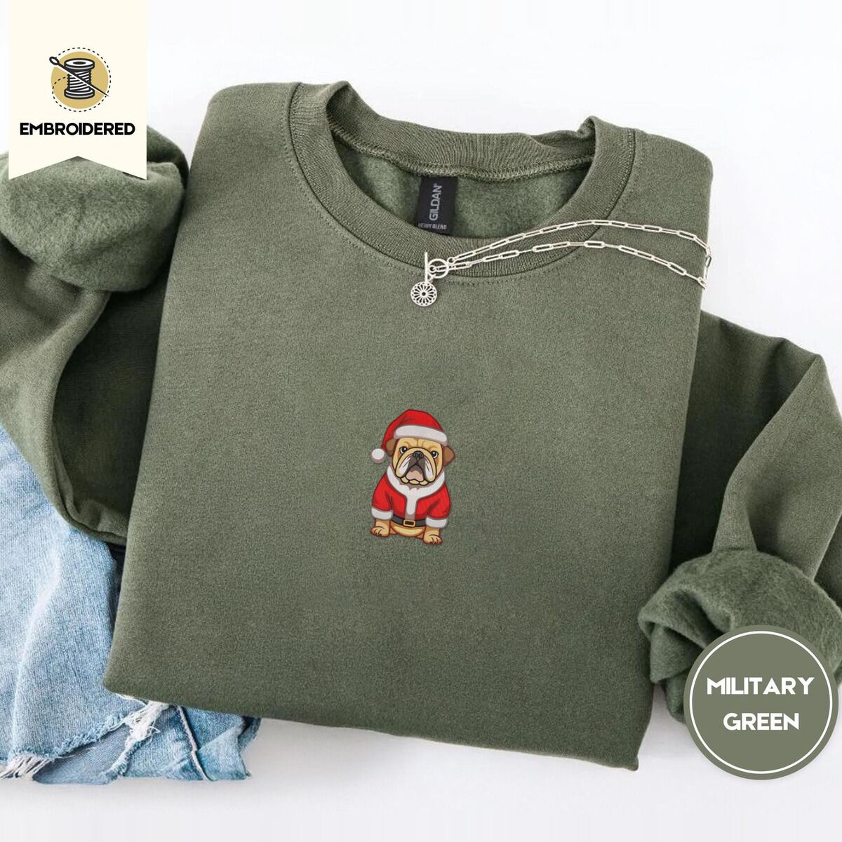English Bulldog Christmas Sweatshirt – Military Green Military Green crewneck sweatshirt folded with light jeans and a silver chain necklace, embroidered with a fawn English bulldog in a red Santa suit, black belt and Santa hat
