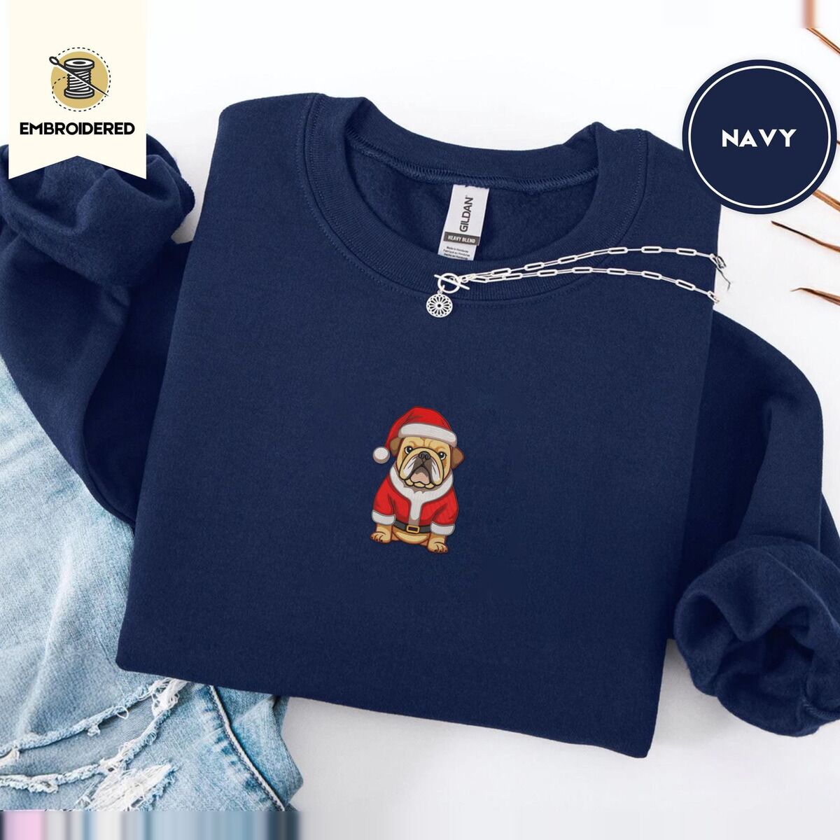 English Bulldog Christmas Sweatshirt – Navy Navy crewneck sweatshirt folded with ripped jeans and a silver chain necklace, embroidered with a fawn English bulldog in a red Santa suit, black belt and Santa hat