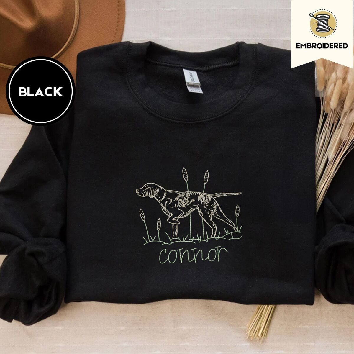 English Pointer Sweatshirt – Black Black crewneck sweatshirt with a brown felt hat and dried bunny tail grass, embroidered with a liver and white English pointer on point among cattails and grass, with an optional name stitched below