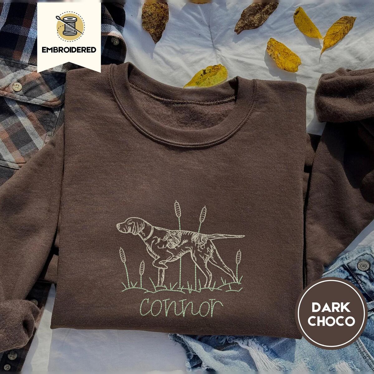 English Pointer Sweatshirt – Dark Chocolate Dark Chocolate crewneck sweatshirt on a plaid flannel shirt with yellow autumn leaves and denim shorts, embroidered with a liver and white English pointer on point among cattails and grass, with an optional name stitched below