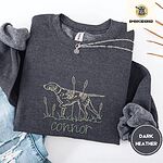 Dark Heather crewneck sweatshirt folded with ripped jeans and a silver chain necklace, embroidered with a liver and white English pointer on point among cattails and grass, with an optional name stitched below