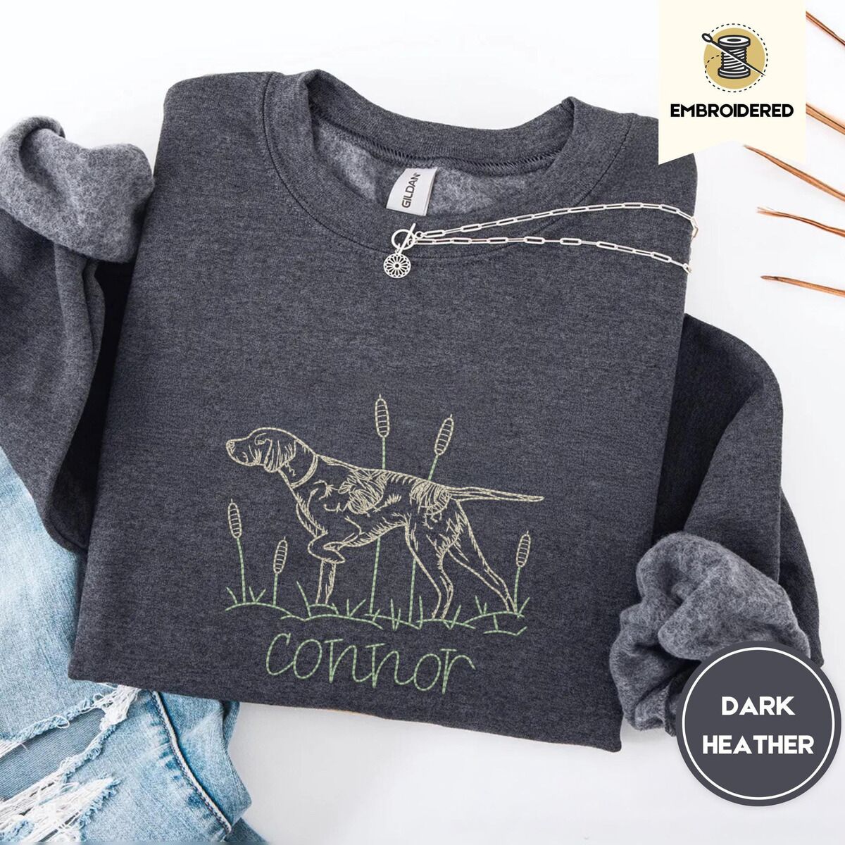 English Pointer Sweatshirt – Dark Heather Dark Heather crewneck sweatshirt folded with ripped jeans and a silver chain necklace, embroidered with a liver and white English pointer on point among cattails and grass, with an optional name stitched below